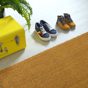 Paillasson - Tapis brosse Coco - Écru - Ep. 23 mm présenté dans une entrée avec chaussures et plante